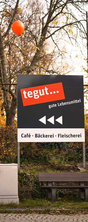 Hinweisschild mit tegut…-Logo am Marktzentrum Ranis mit Aufschrift ‚gute Lebensmittel, Café, Bäckerei, Fleischerei‘, im herbstlichen Außenbereich.