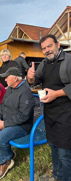 Menschen beim Kürbisfest vor dem Marktzentrum Ranis, Winfried Zein steht mit einem Daumen hoch und einer Tasse in der Hand, während andere Teilnehmer auf einer Bank sitzen. Im Hintergrund ist das Gebäude des Marktzentrums.